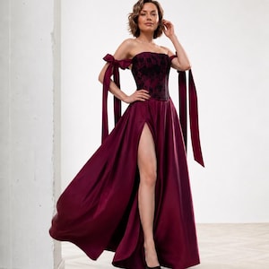 May include: A floor-length burgundy gown featuring a strapless corset-style bodice with a black floral lace overlay. The dress includes off-the-shoulder straps with long, flowing ribbon ties and a high side slit in the satin skirt.