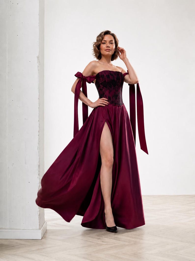 May include: A floor-length burgundy gown featuring a strapless corset-style bodice with a black floral lace overlay. The dress includes off-the-shoulder straps with long, flowing ribbon ties and a high side slit in the satin skirt.