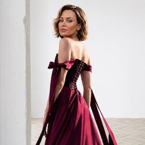 May include: A deep burgundy floor-length gown featuring a strapless corset bodice with a lace-up back closure. The dress includes off-the-shoulder sleeves with long, flowing fabric ties that extend down the sides, creating a dramatic silhouette.