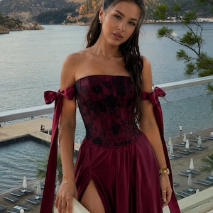 May include: A strapless burgundy gown featuring a corset-style bodice with black floral lace overlay, off-the-shoulder sleeves with long satin ribbon ties, and a floor-length skirt with a high side slit, paired with a small cream-colored handbag.