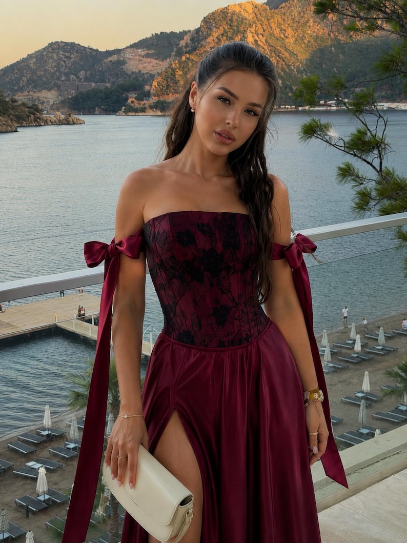 May include: A strapless burgundy gown featuring a corset-style bodice with black floral lace overlay, off-the-shoulder sleeves with long satin ribbon ties, and a floor-length skirt with a high side slit, paired with a small cream-colored handbag.