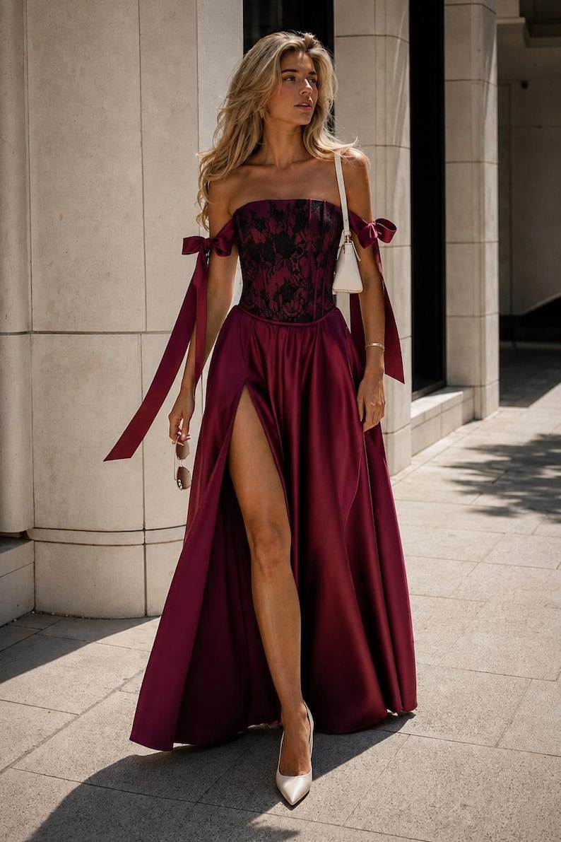 May include: A floor-length burgundy satin gown featuring a strapless corset bodice with black lace overlay, off-the-shoulder ribbon tie sleeves, and a high side slit. The ensemble is paired with a small white shoulder bag and pointed-toe heels.