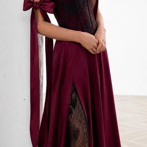 May include: A floor-length burgundy satin gown featuring a strapless corset bodice with black lace overlay, off-the-shoulder sleeves with long satin ribbon ties, and a high side slit revealing a black lace underskirt layer beneath the fabric.