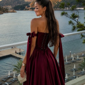 May include: A floor-length, deep burgundy satin gown featuring an off-the-shoulder neckline with tied ribbon straps, a corset-style bodice with lace-up back detailing, and a full, flowing skirt. The dress is a solid, dark red color.
