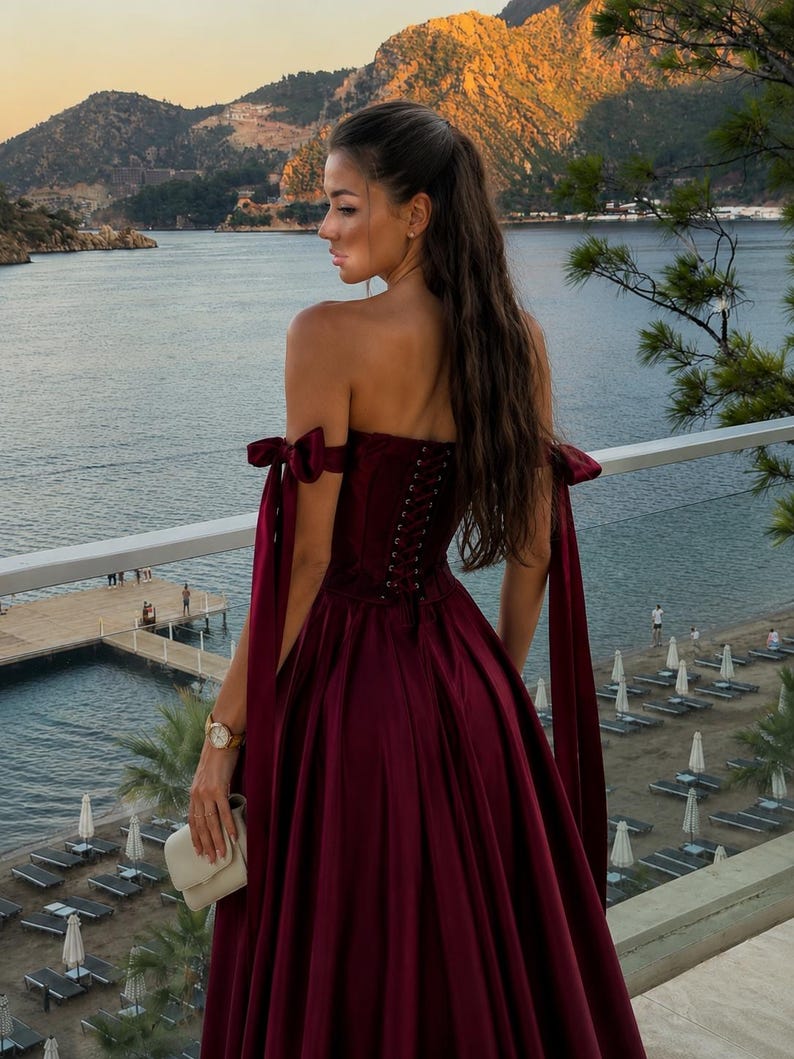 May include: A floor-length, deep burgundy satin gown featuring an off-the-shoulder neckline with tied ribbon straps, a corset-style bodice with lace-up back detailing, and a full, flowing skirt. The dress is a solid, dark red color.