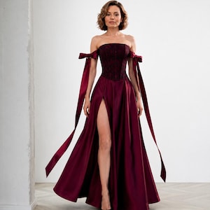 May include: A floor-length burgundy gown featuring a strapless corset bodice with a dark floral pattern, off-the-shoulder sleeves with long ribbon ties, and a full skirt with a high front slit that reveals the leg from the thigh down to the floor.