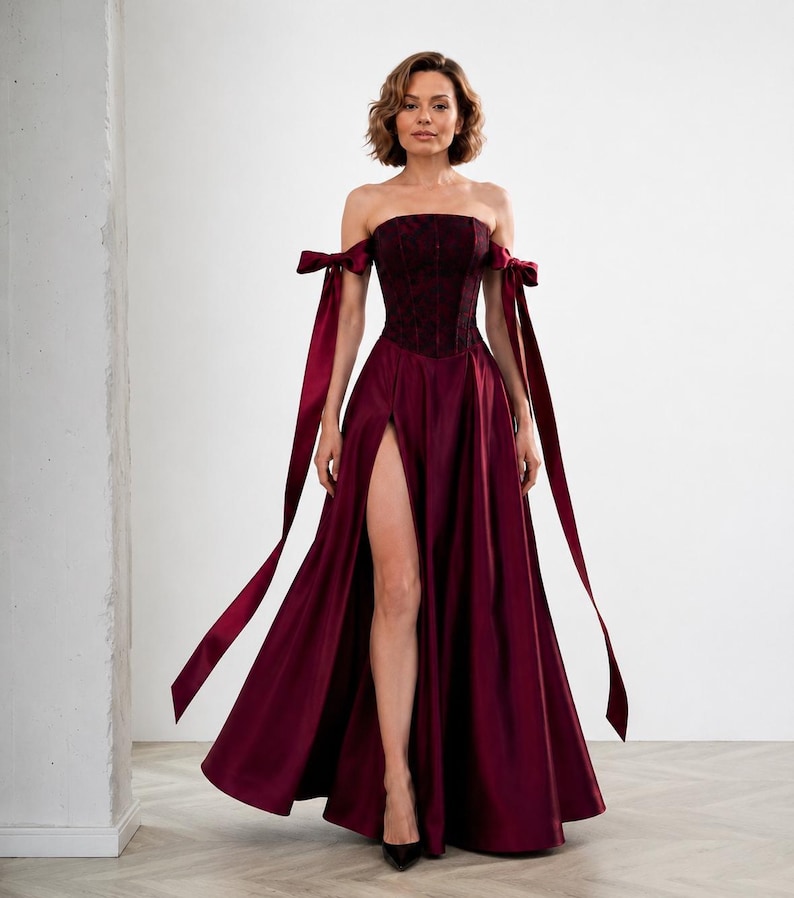 May include: A floor-length burgundy gown featuring a strapless corset bodice with a dark floral pattern, off-the-shoulder sleeves with long ribbon ties, and a full skirt with a high front slit that reveals the leg from the thigh down to the floor.