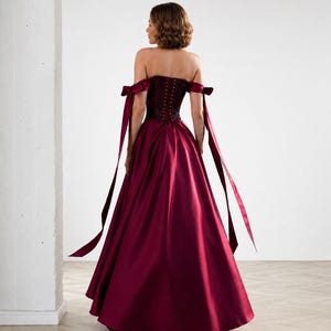 May include: A floor-length burgundy gown featuring a strapless corset bodice with a lace-up back closure. The dress includes off-the-shoulder sleeves with long, flowing ribbon ties that extend down the sides of the full, voluminous satin skirt.