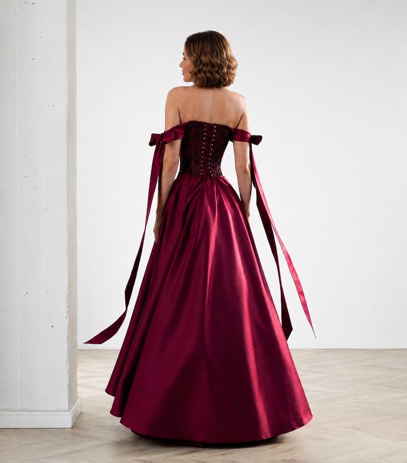 May include: A floor-length burgundy gown featuring a strapless corset bodice with a lace-up back closure. The dress includes off-the-shoulder sleeves with long, flowing ribbon ties that extend down the sides of the full, voluminous satin skirt.