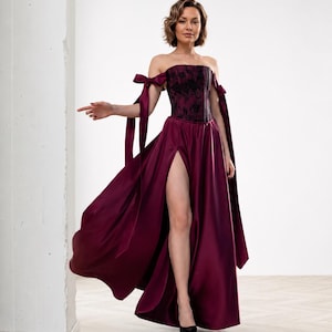 May include: A floor-length burgundy satin gown featuring a strapless corset bodice with black floral lace overlay. The dress includes off-the-shoulder sleeves with long ribbon ties and a high thigh-high slit on the front of the full skirt.