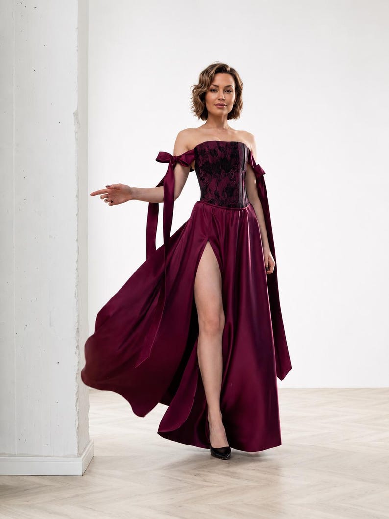 May include: A floor-length burgundy satin gown featuring a strapless corset bodice with black floral lace overlay. The dress includes off-the-shoulder sleeves with long ribbon ties and a high thigh-high slit on the front of the full skirt.