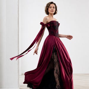 May include: A floor-length gown featuring a deep burgundy satin skirt with a high front slit, revealing a black lace underskirt. The bodice is a structured burgundy and black corset with off-the-shoulder sleeves accented by long satin ribbons.