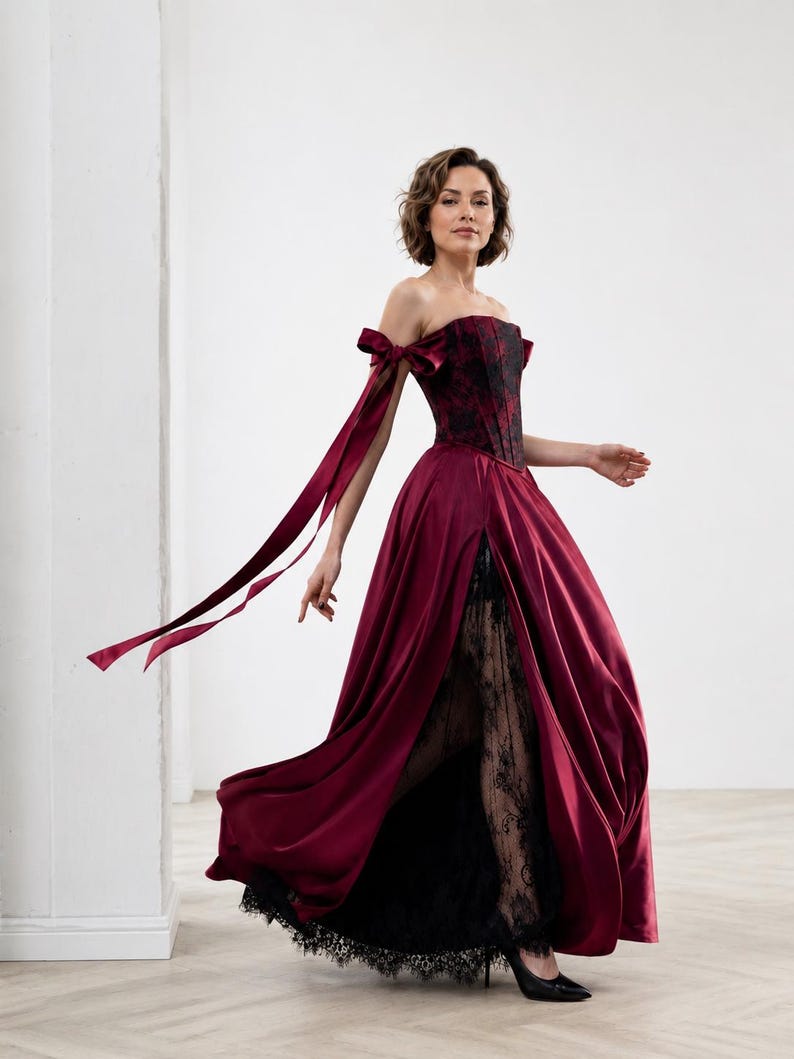 May include: A floor-length gown featuring a deep burgundy satin skirt with a high front slit, revealing a black lace underskirt. The bodice is a structured burgundy and black corset with off-the-shoulder sleeves accented by long satin ribbons.