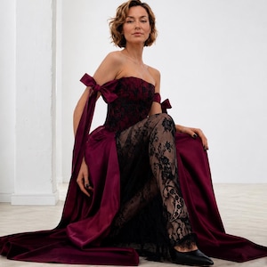 May include: A strapless floor-length gown in deep burgundy satin and black lace. The bodice features a black lace overlay, while the skirt has a satin base with a sheer black lace panel. It includes off-the-shoulder straps with tied bows.