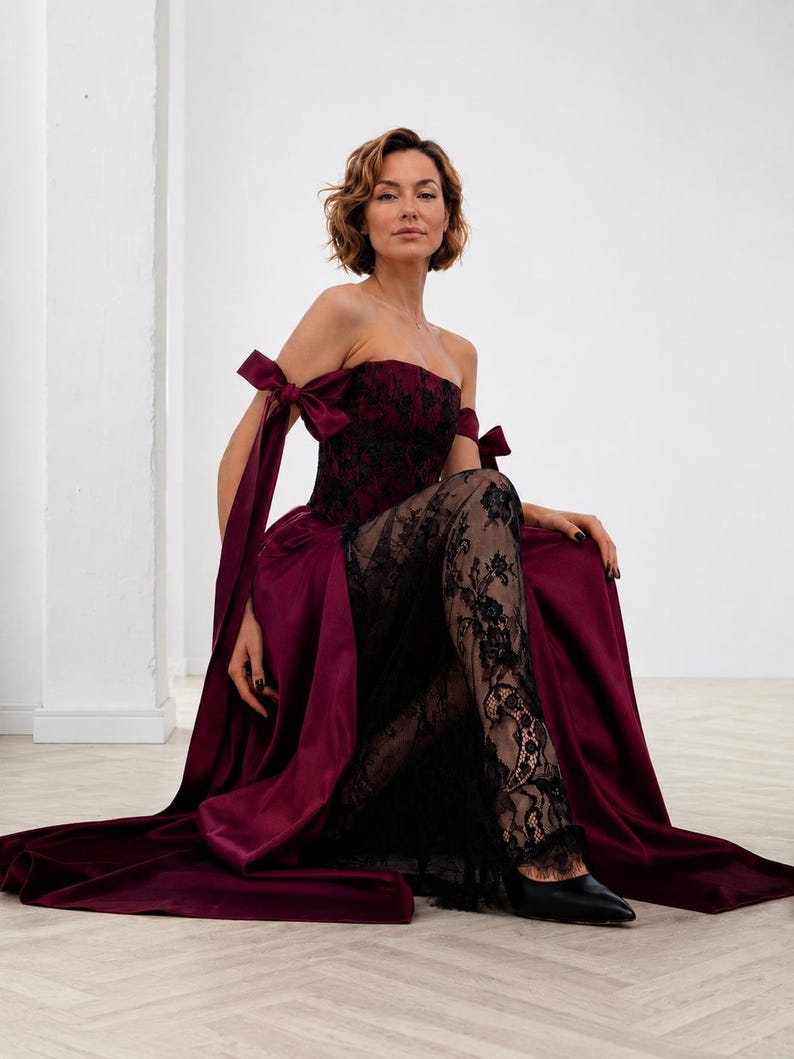 May include: A strapless floor-length gown in deep burgundy satin and black lace. The bodice features a black lace overlay, while the skirt has a satin base with a sheer black lace panel. It includes off-the-shoulder straps with tied bows.
