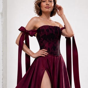 May include: A strapless burgundy gown featuring a corset-style bodice with a black floral pattern and long satin ribbon ties on the shoulders. The skirt includes a high thigh-high slit, creating a flowing silhouette with a deep red satin finish.