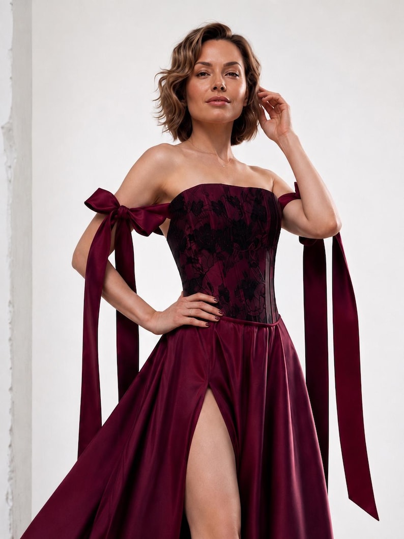 May include: A strapless burgundy gown featuring a corset-style bodice with a black floral pattern and long satin ribbon ties on the shoulders. The skirt includes a high thigh-high slit, creating a flowing silhouette with a deep red satin finish.