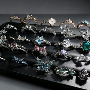 May include: A collection of silver-toned metal rings displayed on a black stand. The rings feature various designs including butterflies, moons, stars, and floral shapes, accented with clear, blue, pink, and green rhinestones and iridescent stones.