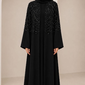 Puede incluir: Una abaya negra hasta el suelo con diseño frontal abierto y mangas largas. La parte superior del pecho y los hombros están adornados con intrincados abalorios negros dispuestos en un delicado patrón de red en cascada. Se lleva un hijab negro a juego.