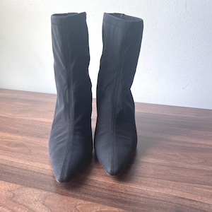 May include: A pair of black ankle boots featuring a pointed toe and a fitted, sock-like shaft. The boots are constructed from a smooth, solid black fabric with visible vertical seam detailing running down the center of the front of each boot.