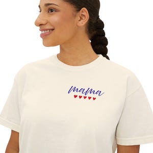 May include: A cream-colored short-sleeve t-shirt featuring the word mama written in a blue cursive script font on the upper left chest area, positioned directly above a horizontal row of five small, solid red heart icons.