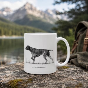Könnte beinhalten: Ein weißer Keramikbecher mit einer schwarz-weißen Illustration eines Deutsch Kurzhaar Hundes im Profil. Der Text DEUTSCH KURZHAAR ist in schwarzen Großbuchstaben unter der Hundeillustration mit einer horizontalen Linie aufgedruckt.