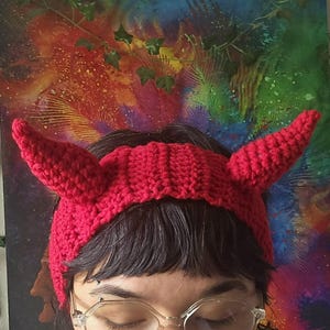 May include: A bright red crocheted headband featuring two prominent, pointed devil-style horns extending from the sides. The headband is constructed with a ribbed texture and is designed to be worn around the head, showcasing a vibrant solid red yarn.