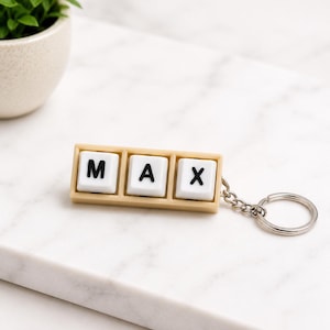 May include: A keychain featuring three white square tiles with the black letters M, A, and X printed on them. The tiles are held within a light tan rectangular frame, which is attached to a silver metal chain and a circular split ring.