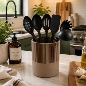 May include: A cylindrical kitchen utensil holder with a ribbed upper section and a smooth base, containing a set of five black cooking tools including a slotted spoon, a solid spoon, a slotted turner, a spatula, and a pasta fork.