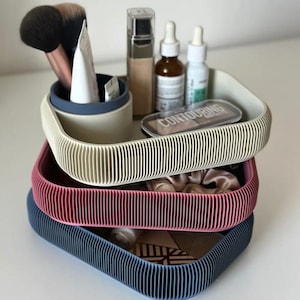 May include: A stack of three square, ribbed storage trays in cream, pink, and blue. The top cream tray contains a blue cylindrical cup holding makeup brushes and tubes, alongside bottles of foundation, serum, and a palette labelled CONTOURING.