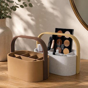 May include: Two oval-shaped plastic storage caddies with arched handles. The front caddy is solid tan with a dark brown handle. The back caddy is speckled light grey with a yellow handle, containing makeup brushes and a NYX contour palette.