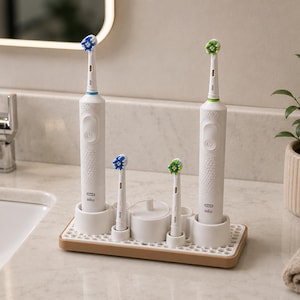 May include: A white bathroom organizer stand with a wooden base and a honeycomb-patterned tray. It holds two electric toothbrushes with white handles, two replacement brush heads, and a small circular container with a lid.