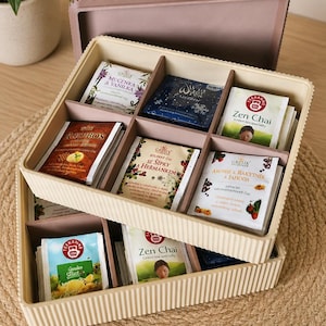 Könnte beinhalten: Eine rechteckige, beige, gerippte Tee-Aufbewahrungsbox mit passendem Deckel, die mehrere Innenfächer enthält, gefüllt mit verschiedenen Teebeuteln, darunter Etiketten wie Zen Chai, Rooibos sowie Aronie & Rakytnik a Jahoda, in einem Raster angeordnet.