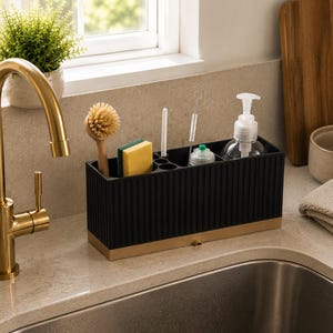 Puede incluir: Un organizador de fregadero rectangular negro con detalles acanalados verticales y una base dorada. El recipiente cuenta con varios compartimentos que contienen un cepillo de madera, una esponja amarilla y verde, un cepillo blanco y un dispensador.