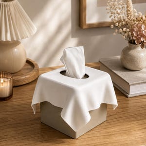 May include: A square tissue box cover featuring a beige base and a white, draped fabric-like top that extends over the sides. A single white tissue is pulled through the central oval opening, standing upright above the smooth, matte surface.