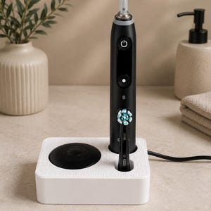 Könnte beinhalten: Eine schwarze elektrische Zahnbürste mit einem weiß-blauen Bürstenkopf steht aufrecht in einer weißen, rechteckigen Ladestation. Der Griff verfügt über einen Ein-/Ausschalter, ein digitales Display und das aufgedruckte Oral-B-Logo.