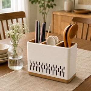 Op de afbeelding: Een rechthoekige witte en beige bestekhouder met drie aparte vakken. De witte buitenkant is voorzien van een horizontale rij verticale uitsparingen voor ventilatie. Het bevat diverse houten lepels, eetstokjes en kleine witte schaaltjes.