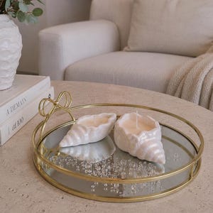 May include: Two pearlescent white seashell-shaped candles with single wicks sit on a round gold-toned metal tray with a mirrored base and a decorative bow-shaped handle. The tray is circular with a thin metal rim and a reflective surface.