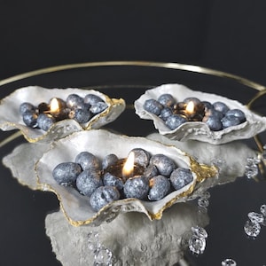 Puede incluir: Tres portavelas de concha de ostra con bordes pintados en oro, cada uno con una pequeña vela encendida rodeada de piedras decorativas de color plata metálica. Las conchas están dispuestas sobre una superficie reflectante con cristales.