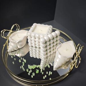 Op de afbeelding: Een rond gouden spiegeldienblad met strikvormige handvatten bevat drie witte kaarsen. Eén kaars is een vierkant blok met een bubbeltextuur en groene accenten, terwijl de andere twee de vorm hebben van zeeschelpen.