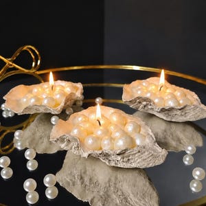 May include: Three lit candles housed in textured, natural oyster shells, each filled with numerous lustrous white pearls. The shells are arranged on a round, reflective gold-toned metal tray featuring a decorative infinity-shaped handle.