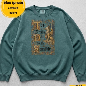 May include: A blue spruce crewneck sweatshirt featuring a graphic print of a griffin and the text 'Tis a Terrible Day to Be Sensible' in an ornate, medieval-style font. The design includes intricate gold and blue scrollwork surrounding the creature.