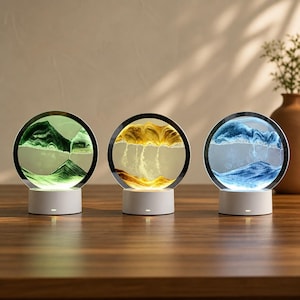 May include: Three circular sand art displays with illuminated bases. Each frame contains liquid and colored sand forming layered landscapes in green, yellow, and blue. The circular glass frames are mounted on white cylindrical stands with lights.