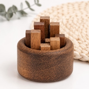 Puede incluir: Un cuenco de madera marrón oscuro que contiene una colección de pequeños bloques rectangulares de madera de diversas alturas y tonos marrones. Los bloques están dispuestos verticalmente dentro del recipiente circular, mostrando texturas de veta natural.