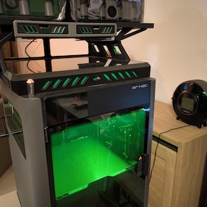 Op de afbeelding: Een Bambu Lab X1C 3D-printer met een groen verlichte printkamer, voorzien van een bovenop gemonteerde AMS-unit met meerdere filamentrollen. De printer heeft een donkergrijs frame, een glazen voordeur en een zwart bedieningspaneel met logo.