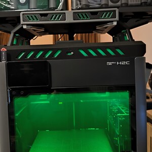 Op de afbeelding: Een zwarte Bambu Lab H2C 3D-printer met een groen getinte glazen voordeur en interne groene verlichting. Het apparaat heeft een rek aan de bovenkant met twee Bambu Lab AMS-units met groene en grijze onderdelen en een zwart bedieningsscherm.