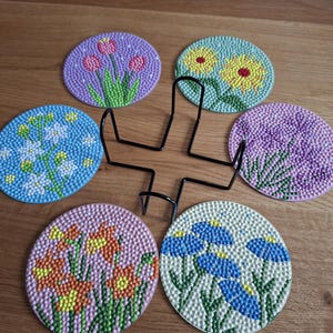 Op de afbeelding: Een set van vijf ronde onderzetters met bloemmotieven in diamond painting stijl in paars, geel, blauw en oranje, gerangschikt rond een zwarte metalen draadhouder. Elke onderzetter toont een uniek bloempatroon gemaakt met kleine, textuurrijke kralen.