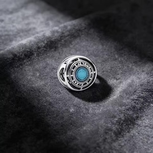 May include: A spherical charm bead featuring a polished silver-tone metal frame with an intricate openwork design. The centre of the bead contains a circular, vibrant cyan blue inlay with a textured, radiating pattern that resembles a glowing core.