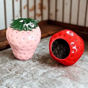 Peut inclure: Un récipient en céramique rose clair en forme de fraise avec un couvercle à feuilles vertes et un bol séparé en forme de fraise rouge contenant des feuilles de thé sombres. Les deux objets ont une finition brillante et texturée.