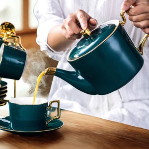 May include: A dark teal ceramic teapot with a gold-toned handle, lid knob, and spout rim, pouring steaming amber liquid into a matching dark teal teacup with a gold-toned handle and rim, set on a matching saucer with a gold-toned edge.